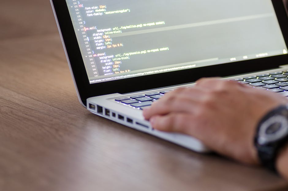 A close-up view of a person's hand operating a laptop with a visible keyboard and a screen displaying lines of colorful code. The laptop is placed on a wooden desk, and the user appears to be engaged 
